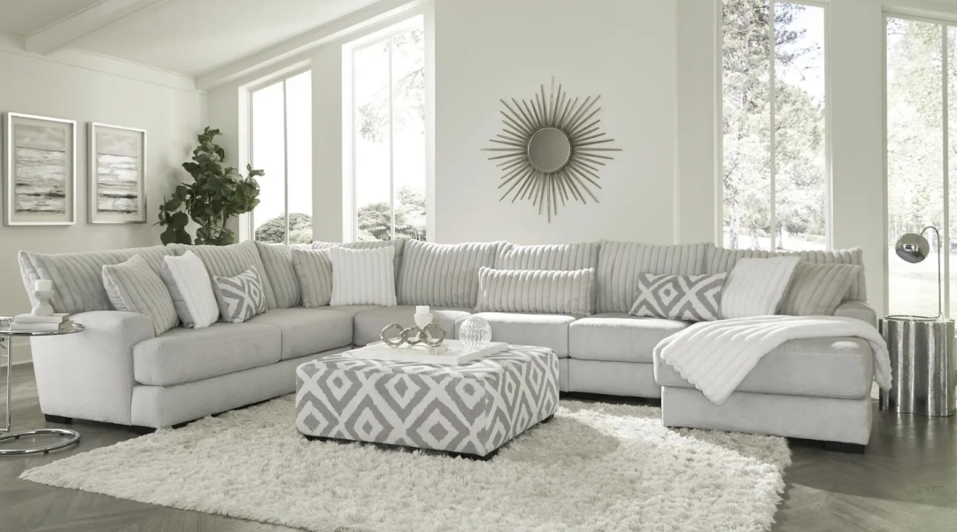 image (1) Modern living room with a large gray L-shaped sofa, geometric-patterned ottoman, fluffy rug, and decorative pillows. A sunburst mirror and several paintings adorn the white walls.