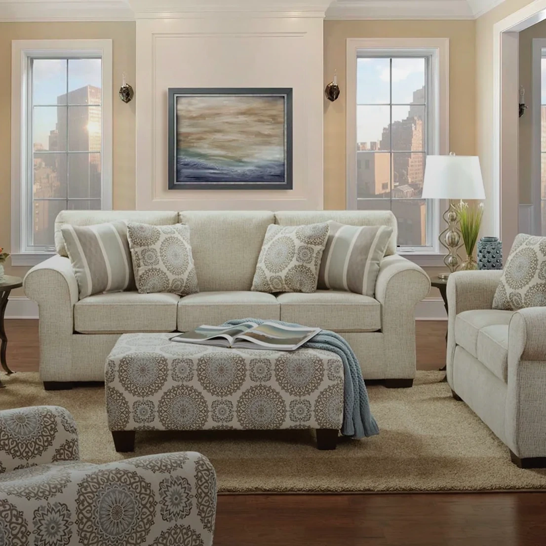 Living room with beige sofas, patterned ottoman, and a landscape painting. Two large windows reveal a cityscape view. A lamp is on a side table next to the sofa.